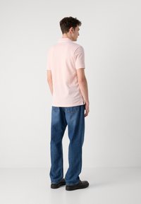 Ανοιχτό ροζ κοντομάνικο πουκάμισο πόλο με κλασικό γιακά, σε συνδυασμό με χαλαρά τζιν μπλε και μαύρα παπούτσια, ιδωμένο από πίσω.