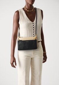 Donna che indossa un top senza maniche a maglia cable color crema e pantaloni, accessorata con una collana in oro, un bracciale e una borsa a tracolla bicolore nera e beige.