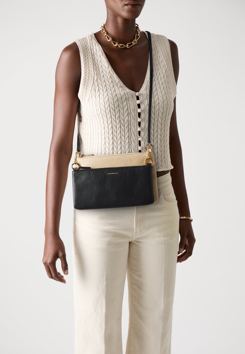 Donna che indossa un top senza maniche a maglia cable color crema e pantaloni, accessorata con una collana in oro, un bracciale e una borsa a tracolla bicolore nera e beige.