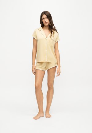 Femme debout pieds nus portant un haut beige à manches courtes avec des boutons et un short assorti, sur un fond blanc uni.