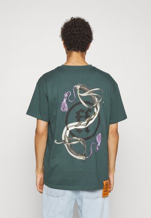 Grøn bomulds t-shirt med et grafisk motiv af sammenflettede sværd med lilla frynser på ryggen. Inkluderer et vævet mærke på siden.