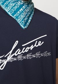 Navy T-shirt with white Lacoste logo; features a patterned turquoise bandana around the neck, showcasing geometric designs.