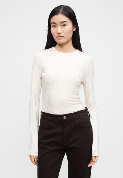 Dolgo rokav, oprijet top v kremni barvi z navpično rebrasto teksturo, kombiniran s temno rjavimi jeans hlačami z zankami za pas in zapiranjem na gumb.