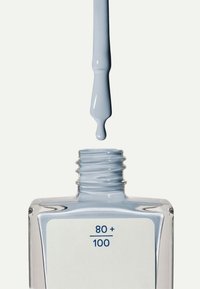 Jasnoniebieski lakier do paznokci kapie z pędzelka nad przezroczystą, prostokątną szklaną butelką z białą etykietą z napisem "80+" i "100".