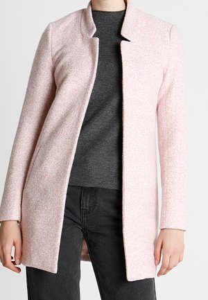 Lyserød uldblandingsfrakke med struktureret finish, lige snit og minimalistisk krave. Båret over en mørkegrå skjorte, kombineret med sorte jeans.