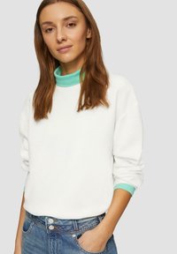 Sudadera blanca con cuello y puños en verde menta, que presenta un corte relajado y textura acanalada, combinada con vaqueros de tiro alto en azul.