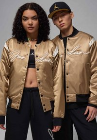 Giubbotti bomber in satin oro con accenti bianchi e scritta "Jordan". Pantaloni sportivi neri; un modello indossa un cappellino nero. Texture morbida, design sportivo.