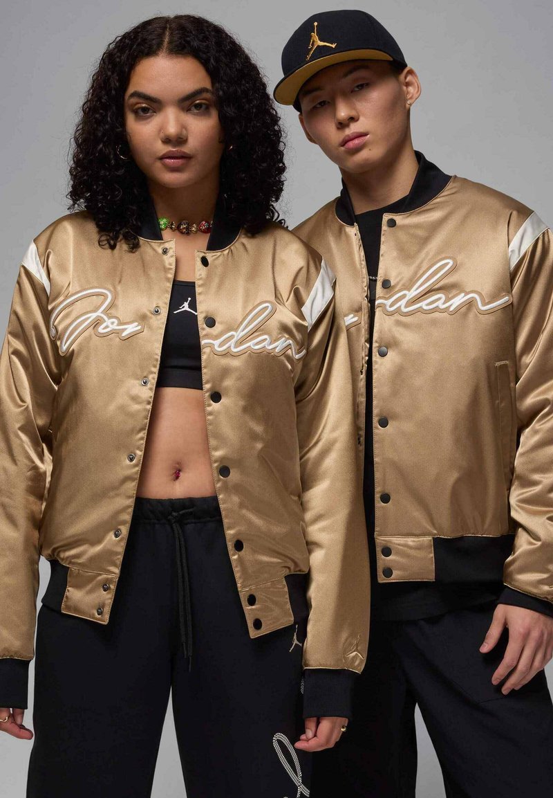 Giubbotti bomber in satin oro con accenti bianchi e scritta "Jordan". Pantaloni sportivi neri; un modello indossa un cappellino nero. Texture morbida, design sportivo.