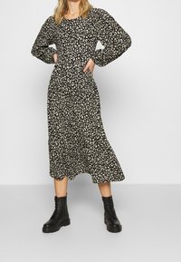 Femme portant une robe midi à manches longues avec un imprimé léopard noir et blanc, associée à des bottes de combat noires à lacets, debout les mains sur les hanches.