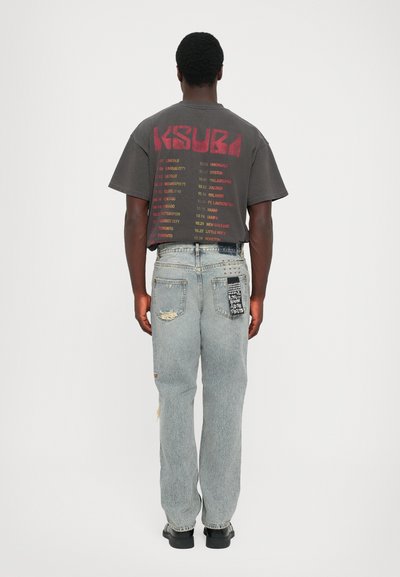 Tricou grafic negru cu text roșu pe spate, purtat cu blugi desenați de culoare albastră deschisă, cu un petic pe șold și uzură vizibilă. Pantofi negri.