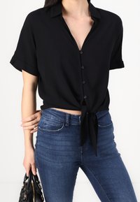 Camisa negra de manga corta con botonadura frontal y detalle de nudo en la cintura, hecha de tela ligera, combinada con jeans ajustados azules.