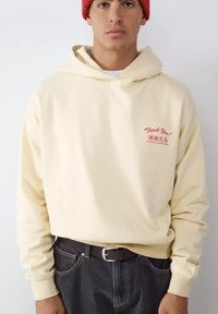 Sudadera con capucha color crema con cordón y puños acanalados, con bordado rojo que dice "Thank You!" en el pecho izquierdo. Se lleva con vaqueros oscuros y un gorro rojo.