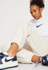 Biely sveter Nike s modrým logom, krémové teplákové nohavice s elastickými manžetami a tmavo-modré a biele tenisky s platformovou podrážkou.
