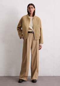 Beige Bomberjacke aus Wolle mit Reißverschluss, geripptem Kragen und zwei Seitentaschen, kombiniert mit beigen weit geschnittenen Hosen und einem cremefarbenen Strickpullover.