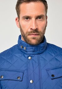 Blå quiltet jakke med høj krave, trykknapper og to forlommer med trykknapper. Glat tekstur med diamantmønster.