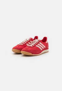 Röda träningsskor i mocka och mesh, med en gummisula, vita snören och tre vita ränder på sidorna.