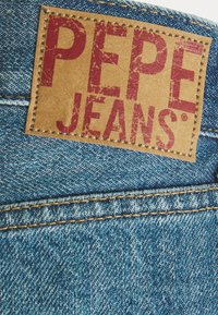 Denim-kangas, jossa on teksturoitu sininen pinta, ja jossa on vaaleanruskea nahkalaatta punaisella "PEPE JEANS" -logolla ja erottuvalla keltaisella ompelella.