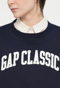 Sudadera navy con "GAP CLASSIC" en bordado blanco en negrita, cuello redondo y superpuesta con una camisa de cuello ligera por debajo.