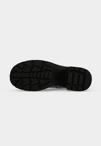 Semelle de botte noire avec des motifs profonds et angulaires pour l’adhérence, présentée à plat sur un fond clair uni.
