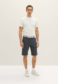 Weißes Kurzarm-T-Shirt kombiniert mit navyblauen gemusterten Shorts. Das Modell steht vor einem hellen Hintergrund und trägt weiße Sneakers.