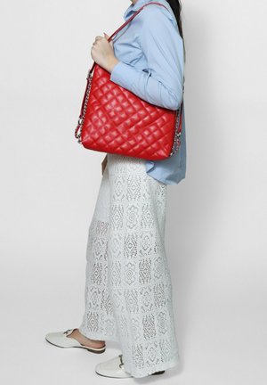 Donna che indossa una camicia azzurro chiaro e pantaloni a gamba larga in pizzo bianco, con una grande borsa trapuntata rossa con tracolla a catena sulla spalla.