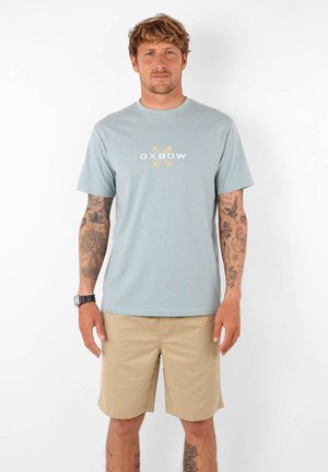 T-shirt en coton bleu clair avec des manches courtes et un col rond. Présente le logo "OXBOW" et des accents floraux en jaune et blanc. Porté avec un short beige.