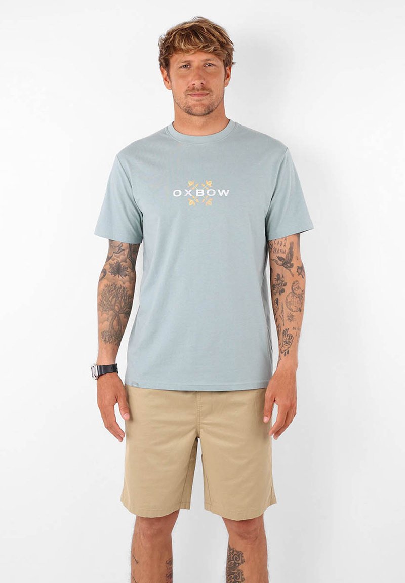 T-shirt en coton bleu clair avec des manches courtes et un col rond. Présente le logo "OXBOW" et des accents floraux en jaune et blanc. Porté avec un short beige.