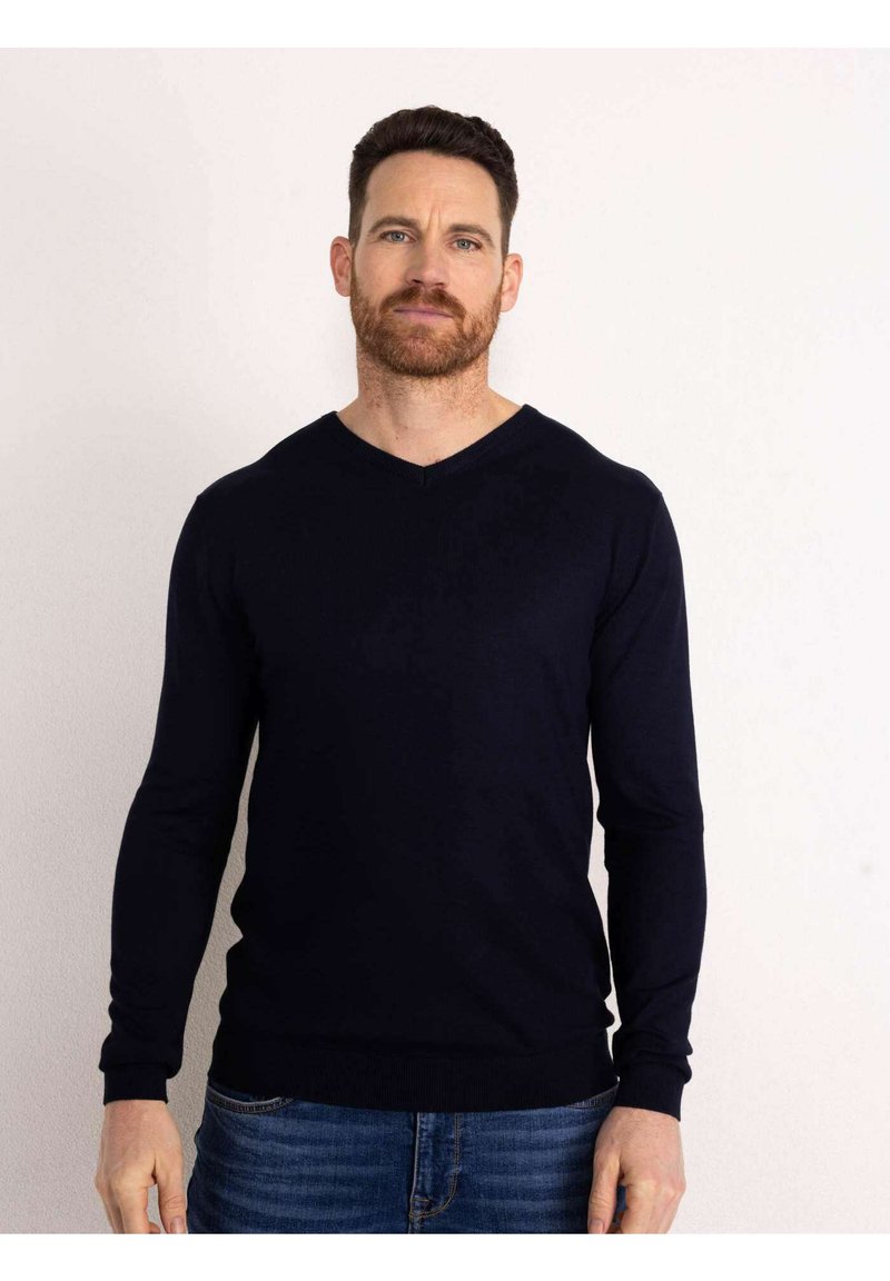 Maglione a V blu navy realizzato in tessuto morbido, con maniche lunghe e orlo aderente, indossato sopra jeans azzurri.