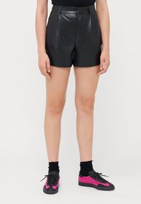 Svarta läderhögmidjade shorts med veck och en figursydd silhuett. Matchas med svarta strumpor och sneakers med rosa accenter.