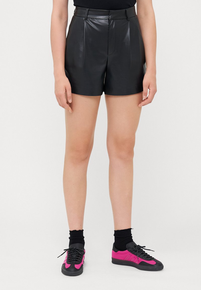 Svarta läderhögmidjade shorts med veck och en figursydd silhuett. Matchas med svarta strumpor och sneakers med rosa accenter.