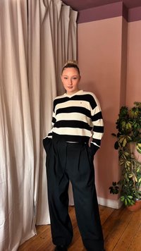 Νεαρή γυναίκα στέκεται μέσα σε εσωτερικό χώρο, φορώντας πουλόβερ με μαύρες και λευκές ρίγες και άνετο μαύρο παντελόνι, με τα χέρια στις τσέπες, και δίπλα της υπάρχει μια γλάστρα με φυτό.