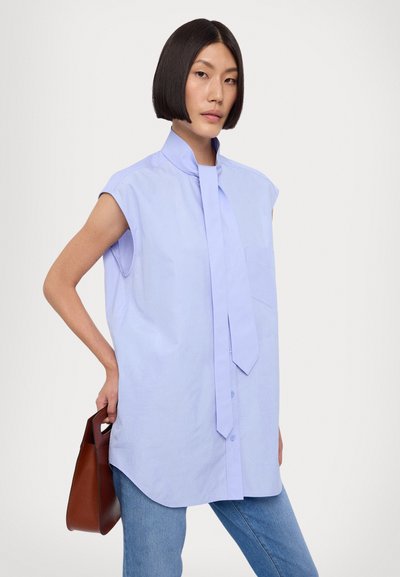 Camicetta senza maniche di un leggero colore lavanda con colletto alto e cravatta. Presenta una tasca e chiusura con bottoni. Abbinata a jeans blu e una borsa marrone.