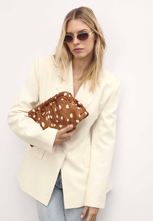 Blazer bianco oversize sopra un top chiaro, che tiene una clutch maculata marrone. Gli occhiali da sole con montatura sottile in oro completano il look.