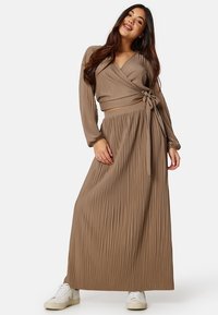 Bruine plissé maxi-jurk met een wikkelontwerp en lange mouwen, met een strik in de taille. Gecombineerd met witte sneakers.