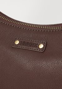Primo piano di una borsa in pelle marrone con texture, dotata di una zip color oro e di un'etichetta rettangolare impressa con la scritta "BRONX" e due borchie dorate.