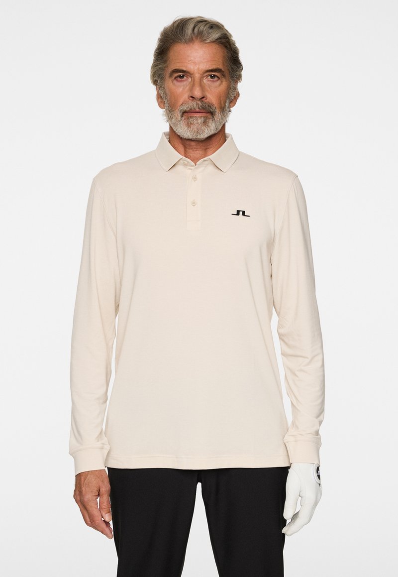Beige Poloshirt mit langen Ärmeln, Kragen und einem schwarzen Logodetail auf der linken Brust. Glatter Stoff mit lockerer Passform.
