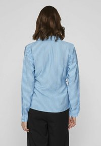 Camisa de rayas azul claro con mangas largas, cuello y detalles de puños con botones. La tela parece suave y tiene un ajuste relajado.