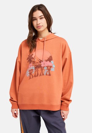 Jeune femme portant un sweat à capuche orange oversize avec un motif floral et palmier tropical et le texte « ROXY », assorti à un pantalon bleu marine avec des rayures orange.