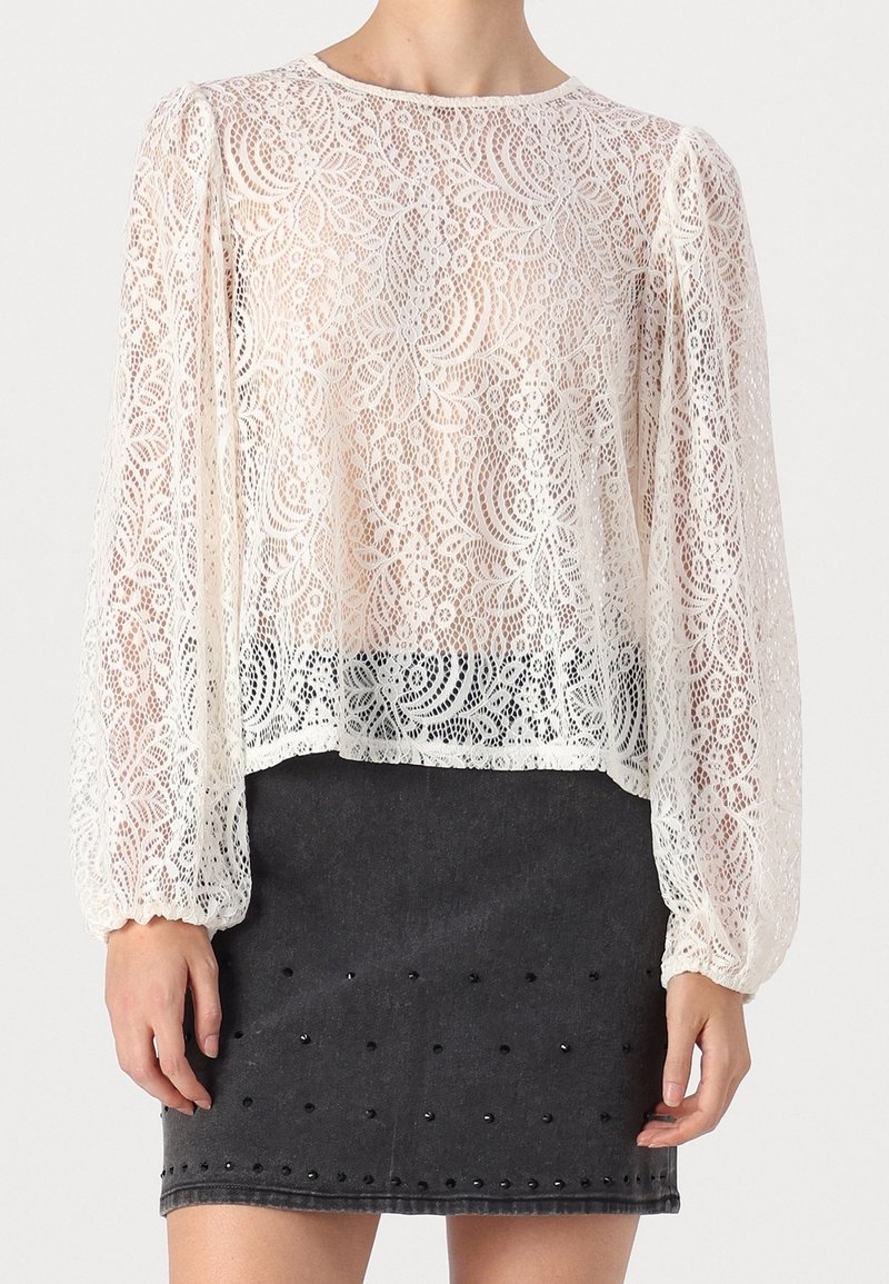 Blouse en dentelle à manches longues bouffantes, présentant des motifs floraux complexes en crème, associée à une jupe en denim foncé ornée de clous noirs.