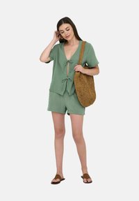 Completo di pantaloni e top in cotone verde con maniche corte, tessuto a trama e un fiocco frontale; abbinato a una borsa a tracolla intrecciata marrone.