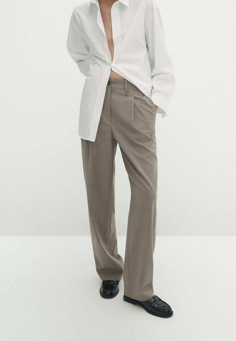 Personne portant un pantalon beige ample, des chaussures noires et une chemise blanche ouverte, une main dans la poche, sur un fond blanc uni.