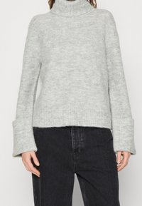 Ljusgrå polotröja med ett texturerat stickmönster, breda ärmmuddar och en kortare passform. I kombination med mörka jeans som lyfter fram outfiten.