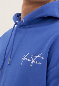 Dettaglio di una felpa blu con cappuccio, con la scritta bianca ricamata "Your Stern" sul petto e i lacci con estremità metalliche sul cappuccio.