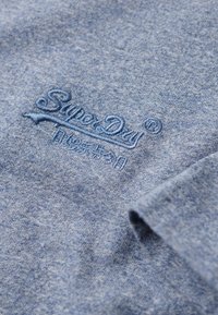 T-shirt en coton bleu avec une texture chinée. Présente un logo "SuperDry" brodé en bleu plus foncé. Design à manches courtes et col rond.