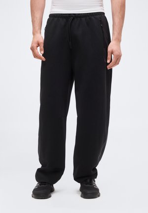 Homme portant un pantalon de survêtement noir ample avec une taille élastique à cordon et des chaussures de sport noires, debout, les bras détendus.