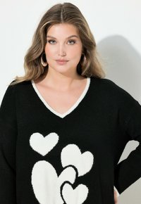 Přerostlý černý svetr s bílými vzory srdcí, s výstřihem do V a kontrastním bílým lemem. Měkká textura a dlouhé rukávy.