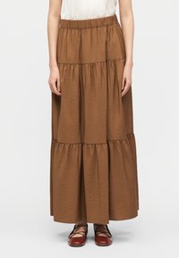 Γυναίκα που φοράει καφέ μακρύ φούστα με βολάν και κόκκινα δερμάτινα επίπεδα παπούτσια με κορδόνια, στέκεται μπροστά σε λευκό φόντο.