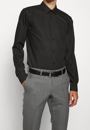 Muž v přiléhavé černé košili s dlouhým rukávem zapnuté na knoflíky, zastrčené do šedých kalhot s černým opaskem, ruce v kapsách.