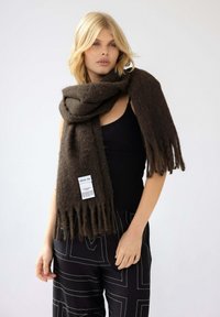Braunfringiger Schal aus weicher Wolle, um den Hals gelegt. Etikett sichtbar mit dem Markennamen "ARCTIC FOX". Schwarzes Oberteil und gemusterte Hose getragen.
