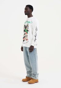 Langärmliges weißes Grafikshirt mit bunten Druckdesigns; lockere hellblaue Jeans; braune Schnürstiefel; silberne Ringe.
